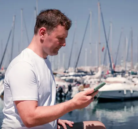 Man in a white t-shirt standing on a marina while looking at his phone